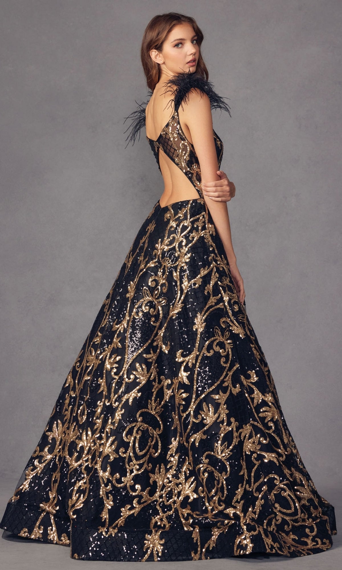 A woman stands against a plain gray background wearing the Feathered-Cap-Sleeve Long Sequin Prom Ball Gown 297, featuring a long black silhouette with gold accents, sequin details, an open back, and feathered cap sleeves.